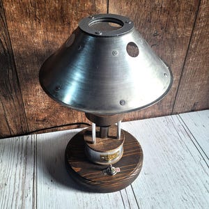 以下が含まれることがあります： 丸い受け皿型のシェードと木製のベースを持つ銀色の金属製のテーブルランプ。シェードには円形の切り抜きが3つと、上部に小さなノブがあります。ベースには、側面に丸い木製のノブがあります。