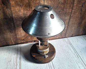 May include: A silver metal table lamp with a round, saucer-shaped shade and a wooden base. The shade has three circular cutouts and a small knob on the top. The base has a round, wooden knob on the side.
