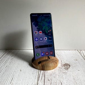 May include: A black smartphone is displayed upright in a wooden phone stand. The stand is round with a cutout design. The phone screen shows app icons. The stand is made of light and dark wood. The background is a white wall and a white wooden surface.