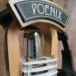 以下が含まれることがあります： 「POENIX」の文字が書かれた黒い看板が付いた木製のヘッドホンアンプのクローズアップ。アンプは透明なガラス管と白と透明のプラスチック部品を備えています。背景には黒いヘッドホンが見えます。
