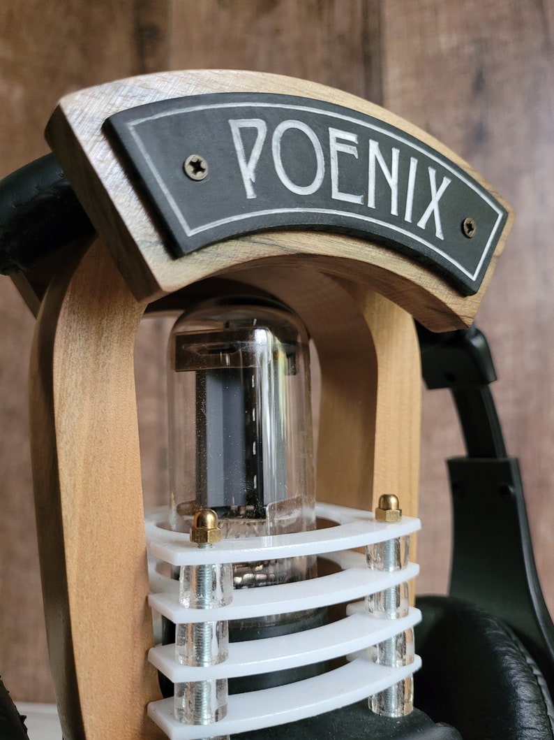 以下が含まれることがあります： 「POENIX」の文字が書かれた黒い看板が付いた木製のヘッドホンアンプのクローズアップ。アンプは透明なガラス管と白と透明のプラスチック部品を備えています。背景には黒いヘッドホンが見えます。