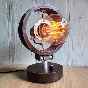 May include: A vintage-style desk lamp with a round, dark brown wooden frame and a large, exposed Edison bulb. The lamp features a circular design with a metal base and a dimmer switch. The warm light from the bulb creates a cozy ambiance.