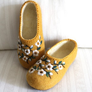 May include: A pair of yellow felt slippers with white embroidered flowers and green leaves. The slippers have a white lining and a stitched edge.