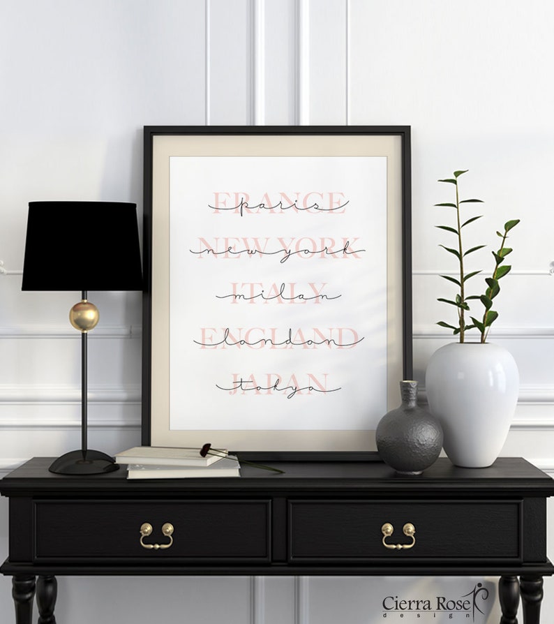 May include: A black framed print with white background and pink text. The text reads "FRANCE paris, NEW YORK, ITALY milan, ENGLAND london, JAPAN tokyo". The print is on a black table with a black lamp and a white vase with a green plant.