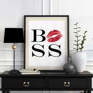 May include: A black and white printable art print with the word "BOSS" in bold black letters. A red lipstick kiss mark is over the "O". The print is framed in a black frame and displayed on a black console table with a black lamp and a vase with green plants.