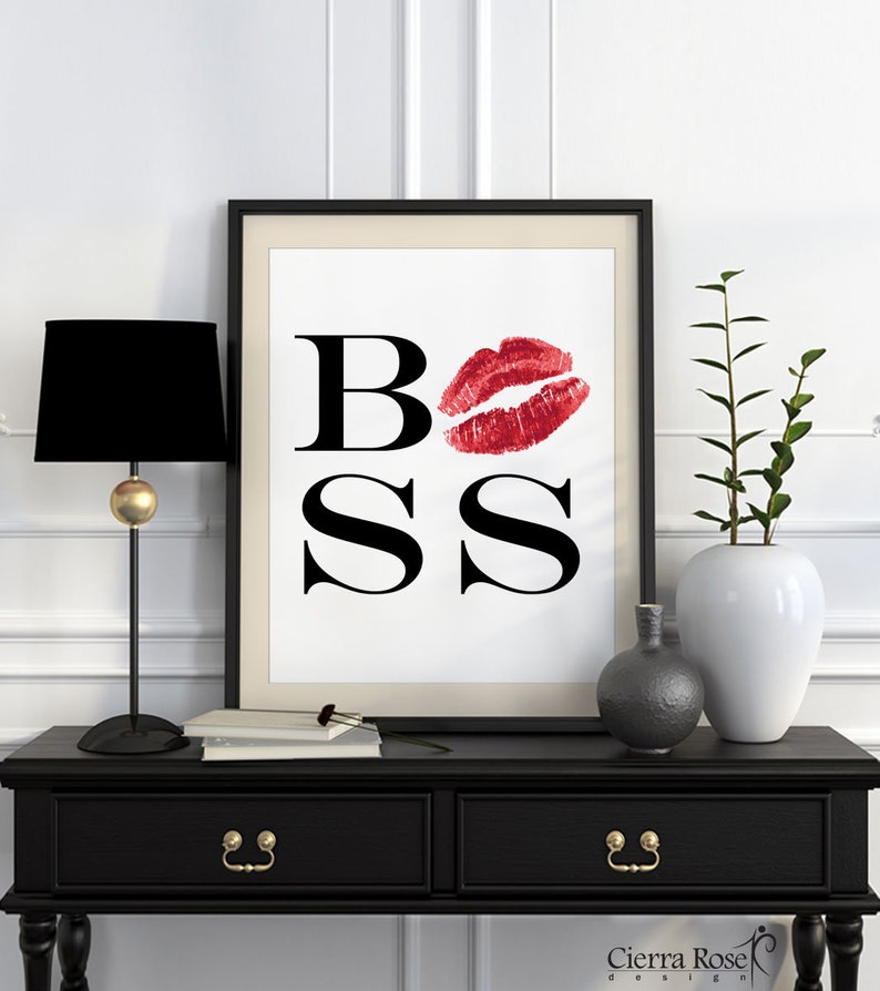 May include: A black and white printable art print with the word "BOSS" in bold black letters. A red lipstick kiss mark is over the "O". The print is framed in a black frame and displayed on a black console table with a black lamp and a vase with green plants.