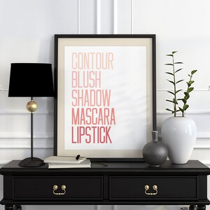 Puede incluir: Una impresi&oacute;n enmarcada en negro con las palabras "CONTOUR BLUSH SHADOW MASCARA LIPSTICK" en un degradado de rosa y rojo. La impresi&oacute;n est&aacute; sobre un fondo blanco y se encuentra sobre una mesa negra con dos cajones. Hay dos jarrones sobre la mesa, uno es blanco y el otro es gris.