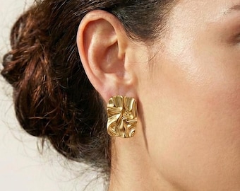 May include: Gold-toned square stud earrings with a textured, rippled surface. The earrings have a unique, abstract design and are shown on an ear. The earrings are a statement piece.