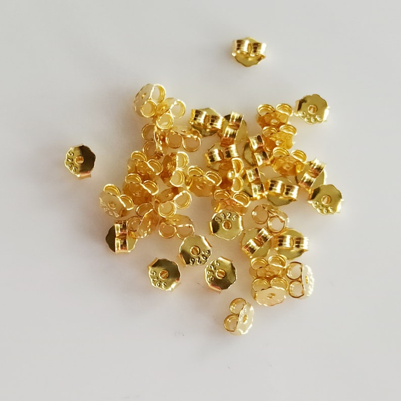 May include: A pile of gold-coloured earring backs. The backs are made of metal and have a small loop on the top. The backs are all the same size and shape.