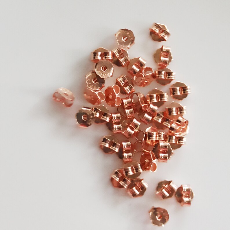 May include: A pile of rose gold earring backs. The backs are made of metal and have a small loop on the back. They are designed to be used with stud earrings.