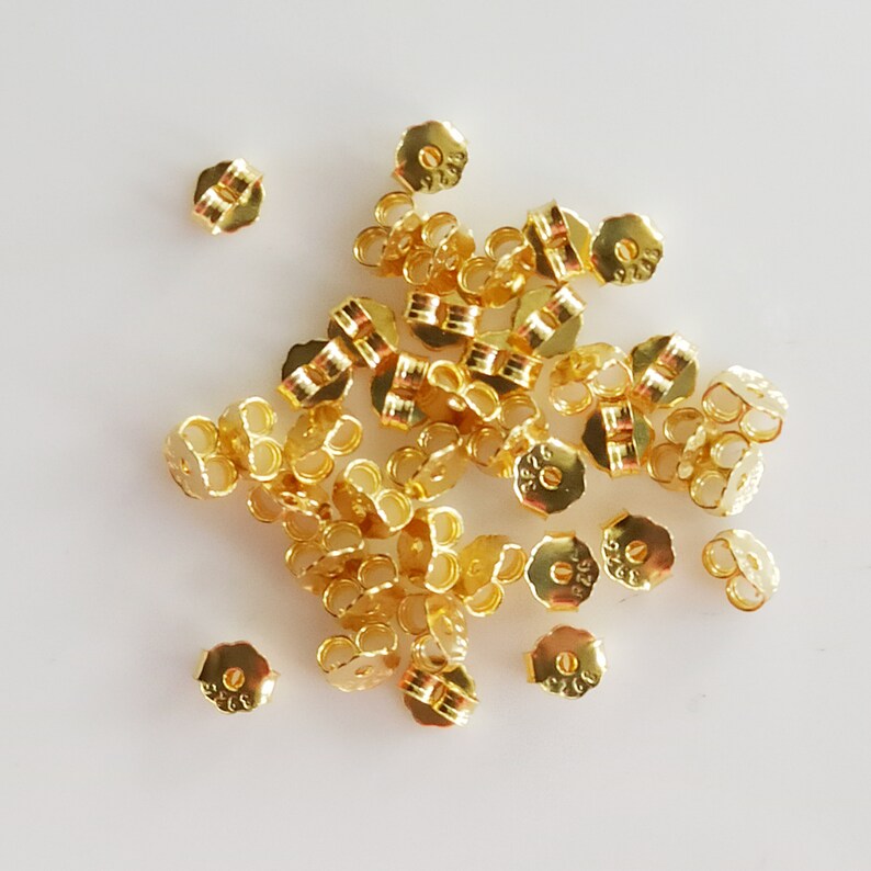 May include: A pile of gold-coloured earring backs. The backs are made of metal and have a small loop on the back. The backs are all the same size and shape.