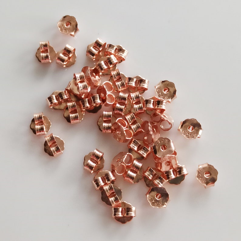 May include: A pile of rose gold earring backs. The backs are made of metal and have a small loop on the top. The backs are all the same size and shape.