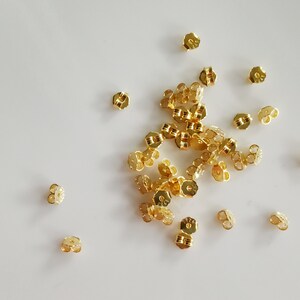 May include: A large group of gold earring backs, scattered on a white surface.