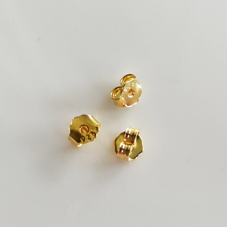 May include: Three gold-coloured earring backs. The backs are designed to be used with post earrings. The backs have a small hole in the centre and a small metal loop on the side.