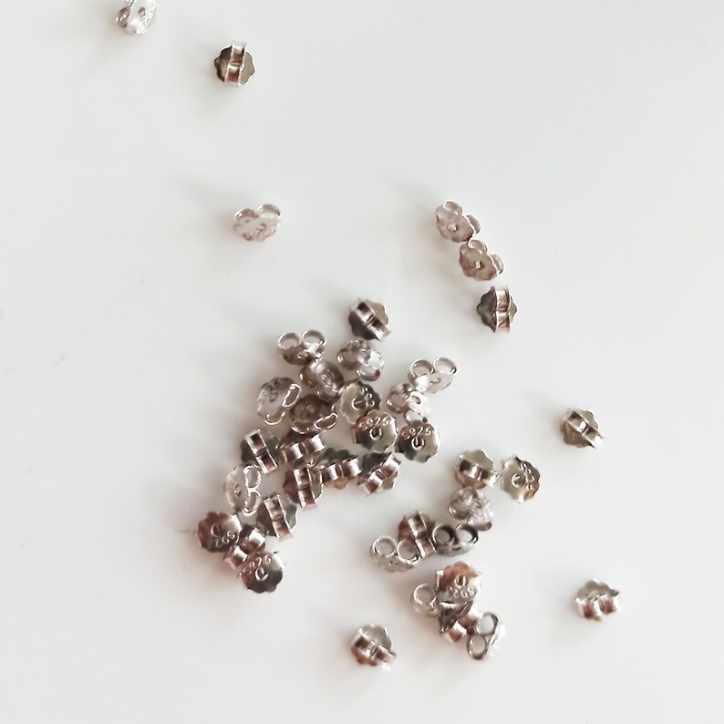 May include: A pile of silver earring backs, some are scattered around the pile. The backs are designed to be used with post earrings.