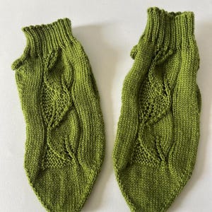 May include: A pair of handmade, green knitted socks. The socks feature a cable knit design with a leaf pattern. The socks are a vibrant shade of green and appear to be made of a soft, warm yarn. The socks are designed to fit the foot.