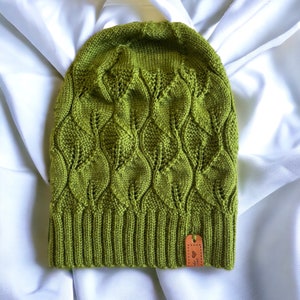 Puede incluir: Un gorro de punto verde con un patrón de hojas. El gorro tiene una etiqueta de cuero marrón con la palabra "Made" en ella.