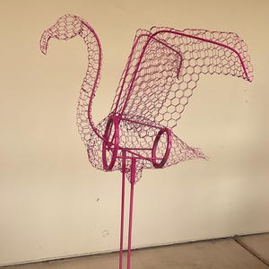 May include: A pink metal flamingo sculpture with a wire mesh body and outstretched wings. The flamingo has a long neck and legs, and is mounted on a square metal base. A decorative garden ornament.