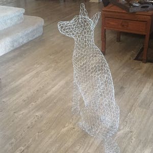May include: A wire mesh sculpture of a dog sitting on a wooden floor. The sculpture is made of chicken wire and is a light gray color.