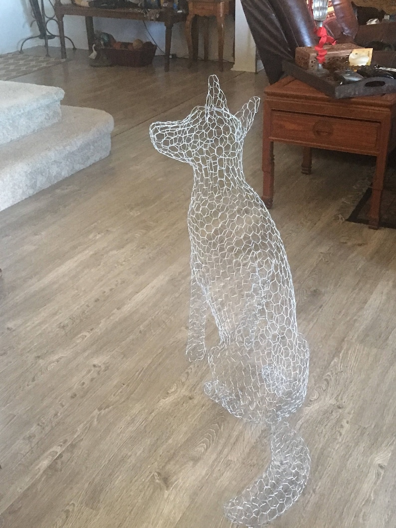 May include: A wire mesh sculpture of a dog sitting on a wooden floor. The sculpture is made of chicken wire and is a light gray color.