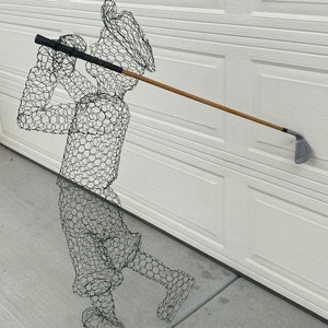 May include: A wire mesh sculpture of a golfer swinging a club. The sculpture is made of black wire and is standing on a grey concrete surface.