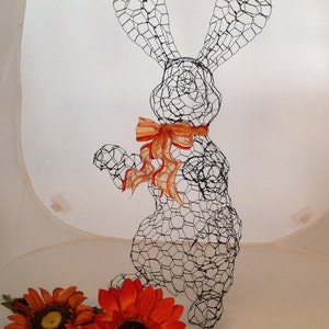 May include: A black wire mesh rabbit sculpture with orange and yellow ribbon around its neck. The rabbit is standing on a white surface with two orange and yellow sunflowers in the foreground.