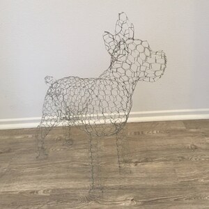 May include: A wire sculpture of a dog, crafted from silver-colored wire. The dog is depicted in a standing pose, with its ears perked up and tail slightly raised. The sculpture is placed on a wooden floor against a white wall.