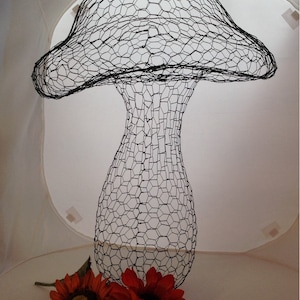 May include: A black wire mesh sculpture of a mushroom with a large cap and a slender stem. The sculpture is sitting on a white surface with two red flowers.
