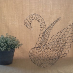 May include: A wire sculpture of a swan with a curved neck and outstretched wing. The swan is made of dark wire mesh. A small blue pot with white flowers sits to the left of the swan. The background is a light beige color.
