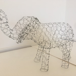May include: A wire mesh sculpture of an elephant, with a long trunk raised in the air. The sculpture is made of a single piece of wire mesh, and the elephant's features are formed by the way the mesh is bent and shaped.