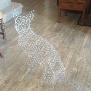 May include: A wire mesh dog sculpture in a sitting position. The sculpture is made of a silver wire mesh and is sitting on a light brown wood floor.