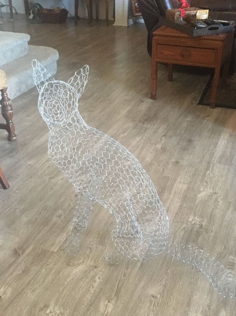 May include: A wire mesh dog sculpture in a sitting position. The sculpture is made of a silver wire mesh and is sitting on a light brown wood floor.