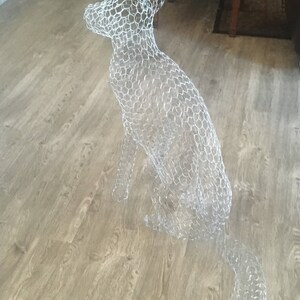 May include: A wire mesh sculpture of a dog sitting on a wooden floor. The sculpture is made of chicken wire and is shaped like a dog.