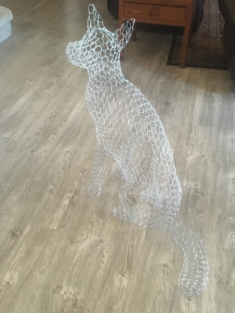 May include: A wire mesh sculpture of a dog sitting on a wooden floor. The sculpture is made of chicken wire and is shaped like a dog.