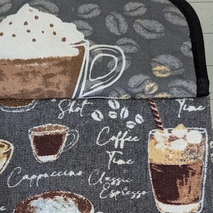 May include: A gray kitchen towel with a coffee theme. The towel features various coffee cups, coffee beans, and the words "Coffee Time", "Cappuccino", and "Espresso". The top portion has a large coffee cup with whipped cream.