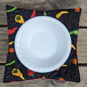 May include: A white bowl sits on a black, square bowl cozy. The cozy is patterned with colorful chili peppers in red, yellow, and green. The cozy has a red border.