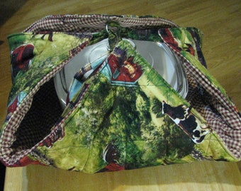 May include: A quilted casserole carrier with a farm scene print in green, red, and brown. The carrier has a gingham lining and a metal ring for carrying. The carrier is designed to hold a round casserole dish.