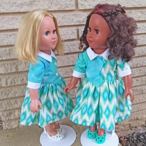 May include: Two dolls wearing matching blue and green dresses with white collars. The dolls have different skin tones and hairstyles. One doll has blonde hair and blue eyes, while the other doll has brown hair and brown eyes.
