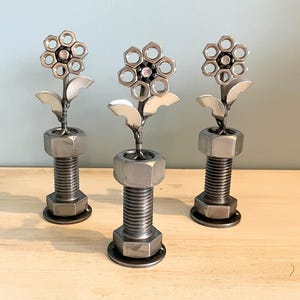 May include: Three metal flower sculptures. Each flower has a circular head made of multiple small, round pieces. The flowers have silver leaves and are mounted on a large bolt base. The background is a light blue wall and a wooden surface.