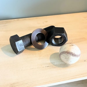 May include: A large metal bolt, nut, and washer, along with a white baseball, are arranged on a light wood surface.