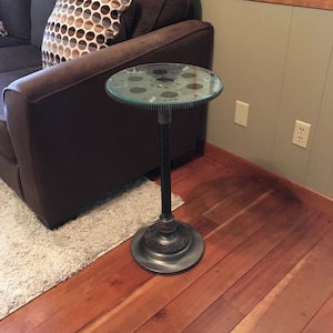 May include: A unique side table with a round glass top supported by a metal gear and a dark metal base. The table stands on a dark metal base and is placed on a hardwood floor. The table is next to a brown sofa and a white rug.