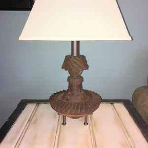 May include: A rustic lamp with a white lampshade and a base made from repurposed metal gears. The lamp is sitting on a white table with black trim.