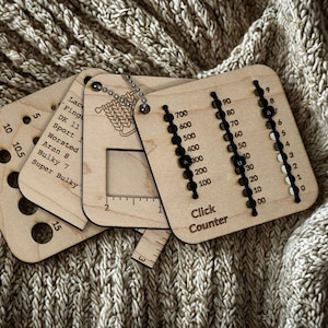May include: Wooden knitting tools, including a click counter and gauge, are displayed. The click counter has black beads and the text "Click Counter". The gauge has various hole sizes and yarn weight labels. The tools are on a textured, neutral-toned surface.