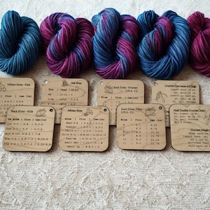 May include: An overhead shot displays several skeins of yarn in shades of blue, purple, and pink, alongside wooden tags with size charts and crochet instructions. The yarn is arranged on a textured, off-white surface, ideal for knitting and crochet projects.