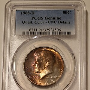 May include: A 1968-D 50 cent half dollar coin graded by PCGS as Genuine, Quest. Color - UNC Details, with a grading number of 6711.91/32924506. The coin is encased in a clear plastic holder.