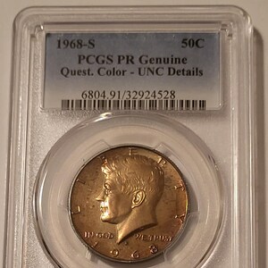 May include: A 1968-S Kennedy half dollar coin, graded by PCGS, encased in a protective holder. The coin features a profile of John F. Kennedy and the text "IN GOD WE TRUST". The holder displays "50C" and "PCGS PR Genuine Quest. Color - UNC Details".