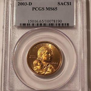 May include: A gold-colored 2003-D Sacagawea dollar coin, encased in a clear plastic holder. The coin features a profile of Sacagawea and her child. The holder has text that reads "2003-D", "PCGS MS65", and "SAC$1".