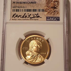 May include: A 2004 S $1 gold-colored coin encapsulated in a clear NGC slab. The coin features the image of Sacagawea with her child, the text LIBERTY, IN GOD WE TRUST, and 2004. The label includes PF 70 ULTRA CAMEO and a signature by Randy'L Teton.