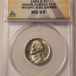 May include: A 1985-D 5-cent coin, graded MS 63, encased in a clear protective holder. The coin features a profile of Thomas Jefferson. The holder displays the coin's weight as 4.82 grams and the text "MINOR CURVED CLIP".