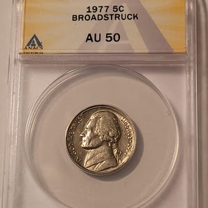May include: A 1977 Jefferson nickel coin encapsulated in a clear plastic ANACS slab. The label reads 1977 5C BROADSTRUCK AU 50. The silver-colored coin features the profile of Thomas Jefferson, the text IN GOD WE TRUST, LIBERTY, and 1977.